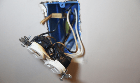 An outlet hganging out of the wall, having clearly sparked or caught fire.